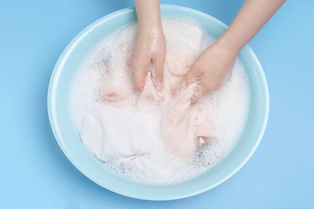 Hands washing clothes in a blue basin filled with soapy water, creating bubbles, against a light blue background.