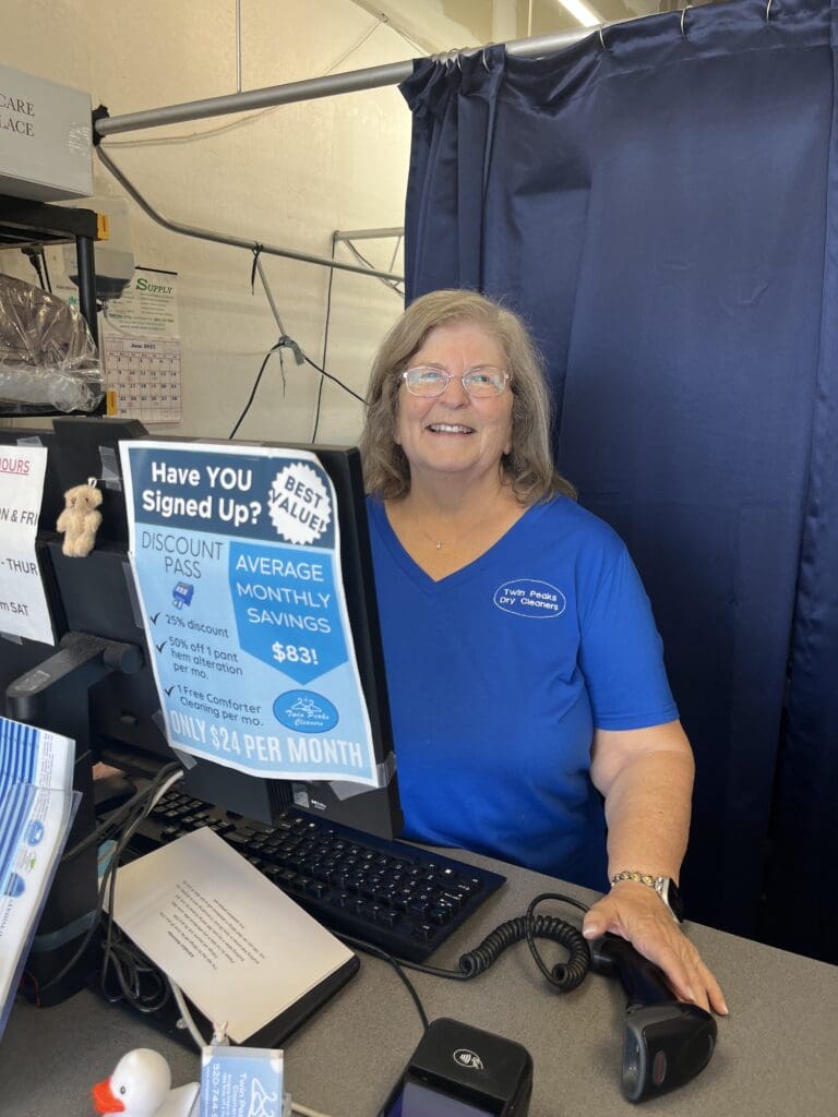 A smiling woman in a blue shirt stands behind a counter at a dry cleaning store, with a computer monitor and a sign promoting a discount pass visible.