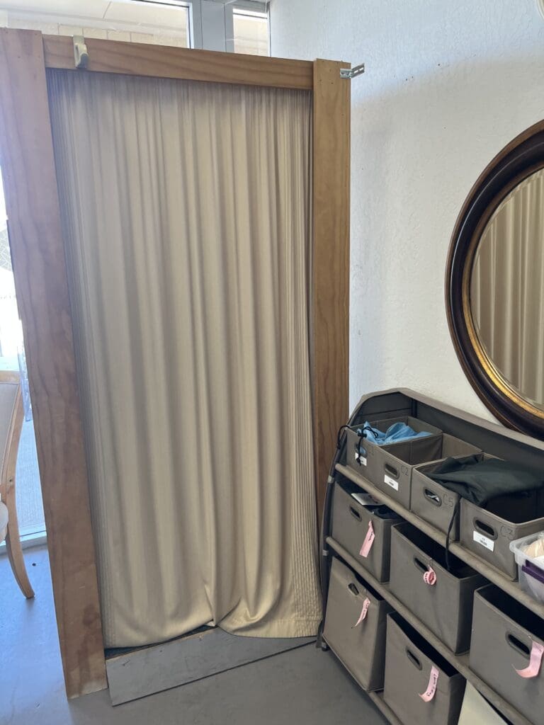 A beige curtain hangs in a wooden frame next to a round mirror and an organizer with multiple fabric storage bins.