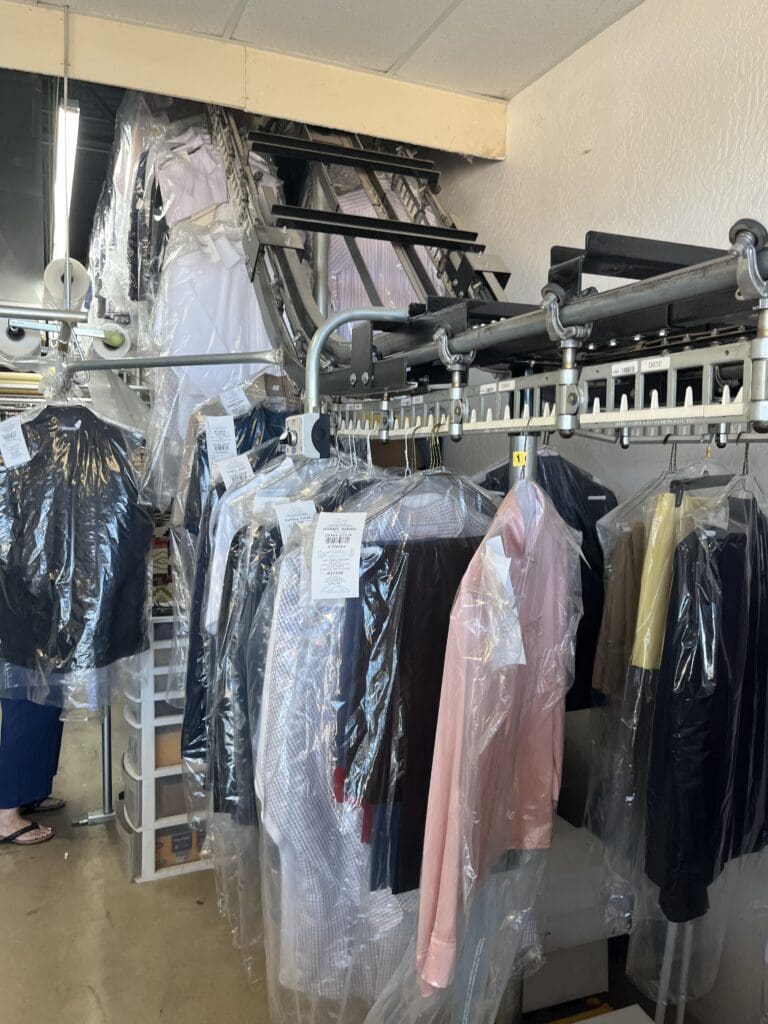 Clothing covered in plastic hangs on a conveyor rack in a dry cleaning shop, with various shirts, jackets, and dresses visible.