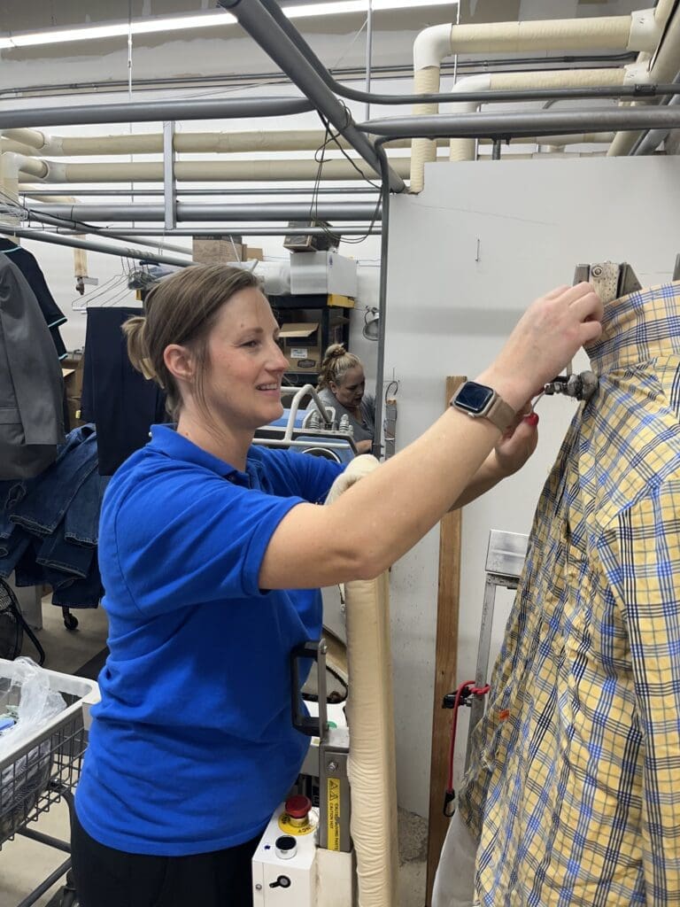A woman in a blue shirt operates a machine while working with a yellow plaid shirt in a laundry or dry cleaning facility.