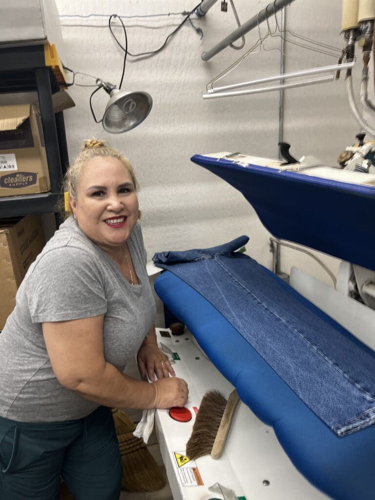 A woman smiles while pressing blue jeans on a large ironing machine in a laundry or dry cleaning workspace.