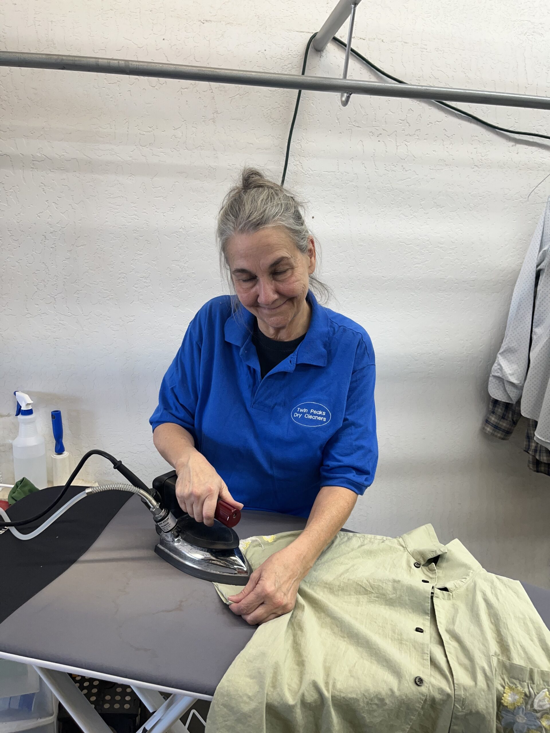A woman in a blue shirt is ironing a light green button-up shirt on an ironing board in a laundry or dry cleaning setting.