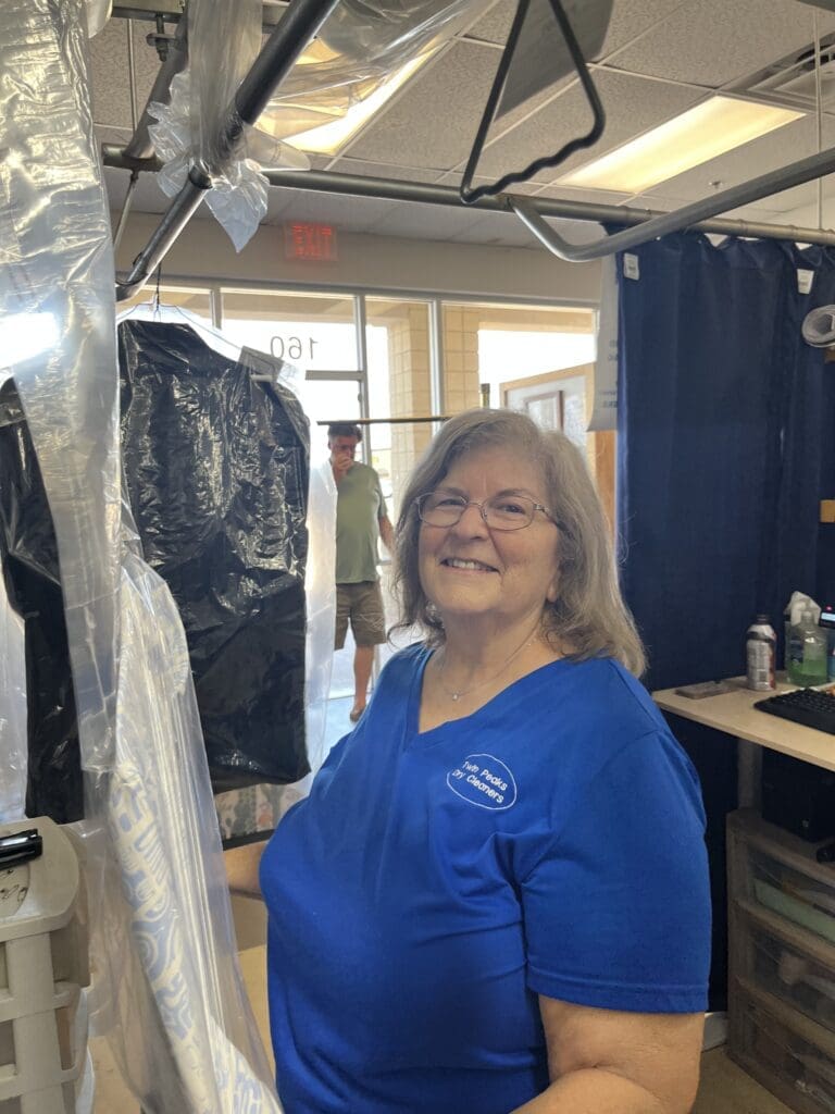 A woman wearing a blue shirt and name tag stands inside a dry cleaning shop, smiling at the camera. Garments covered in plastic hang beside her. A man is visible in the background.