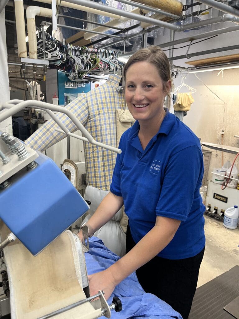A woman in a blue uniform operates a pressing machine at a dry cleaning facility, with clothes hanging on racks in the background.