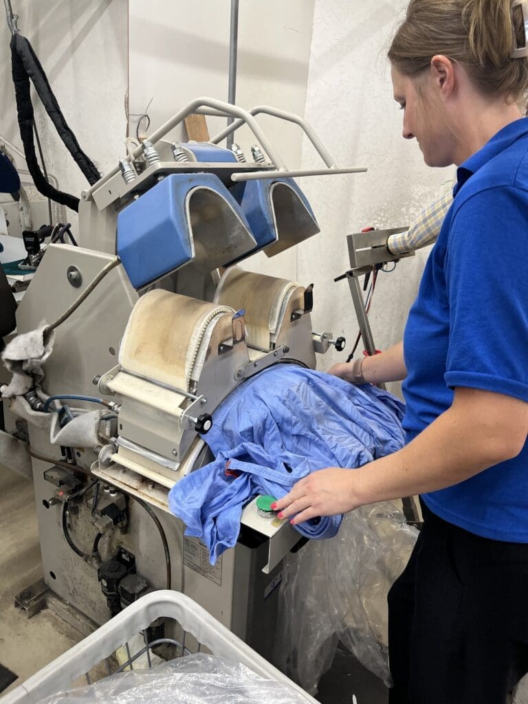 A person operates a commercial ironing press, smoothing a blue garment in an industrial laundry setting.