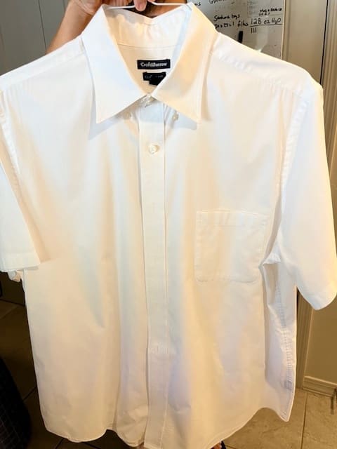 A person holding a short-sleeve, white button-up shirt with a front pocket, displayed indoors.