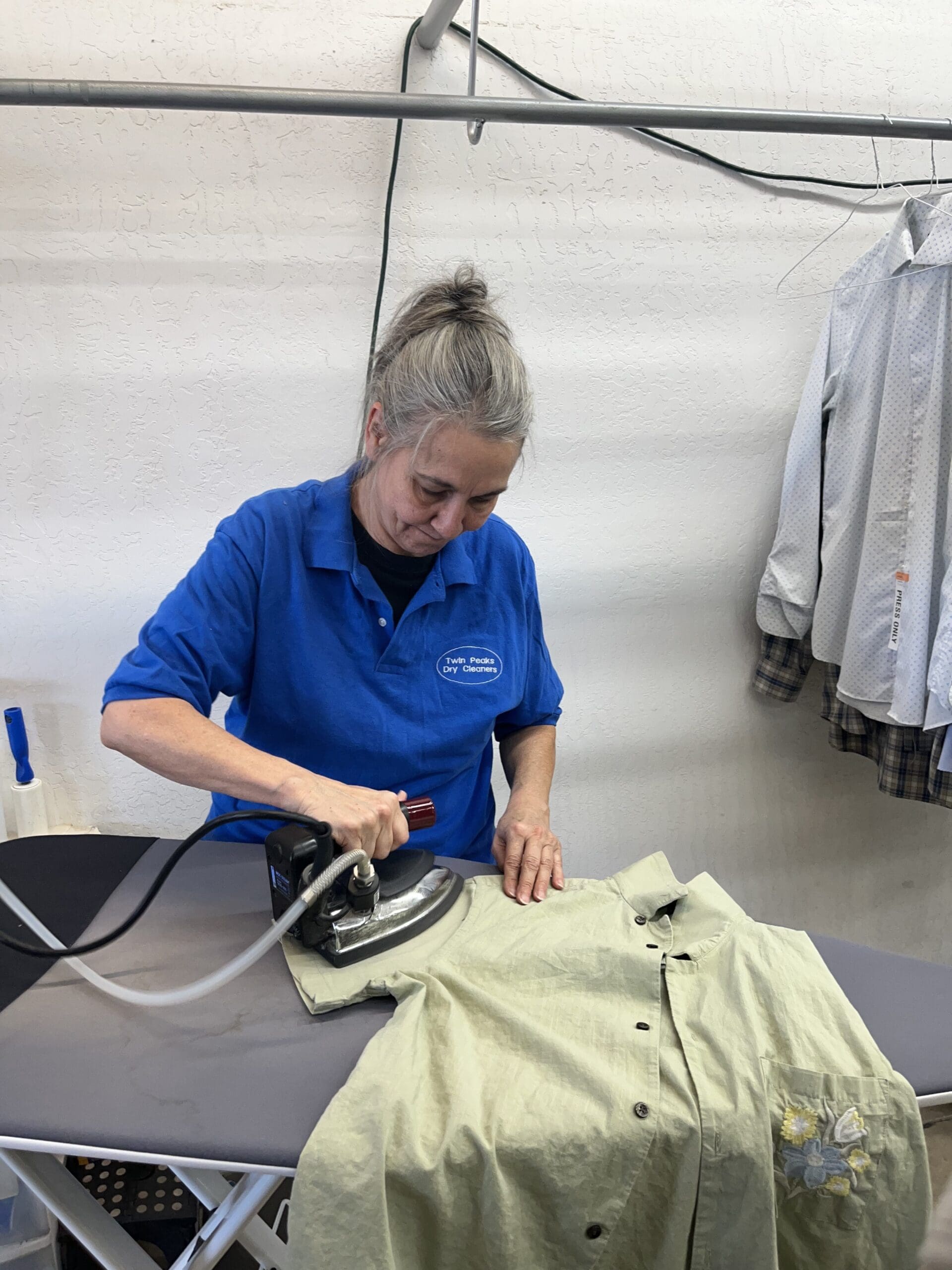 A woman in a blue shirt irons a green button-up shirt on an ironing board; other shirts hang in the background.