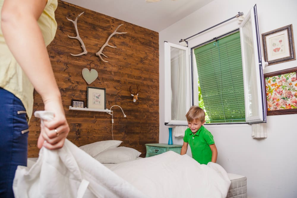 An adult and a young boy make a bed together in a bedroom with a rustic wooden accent wall and an open window.