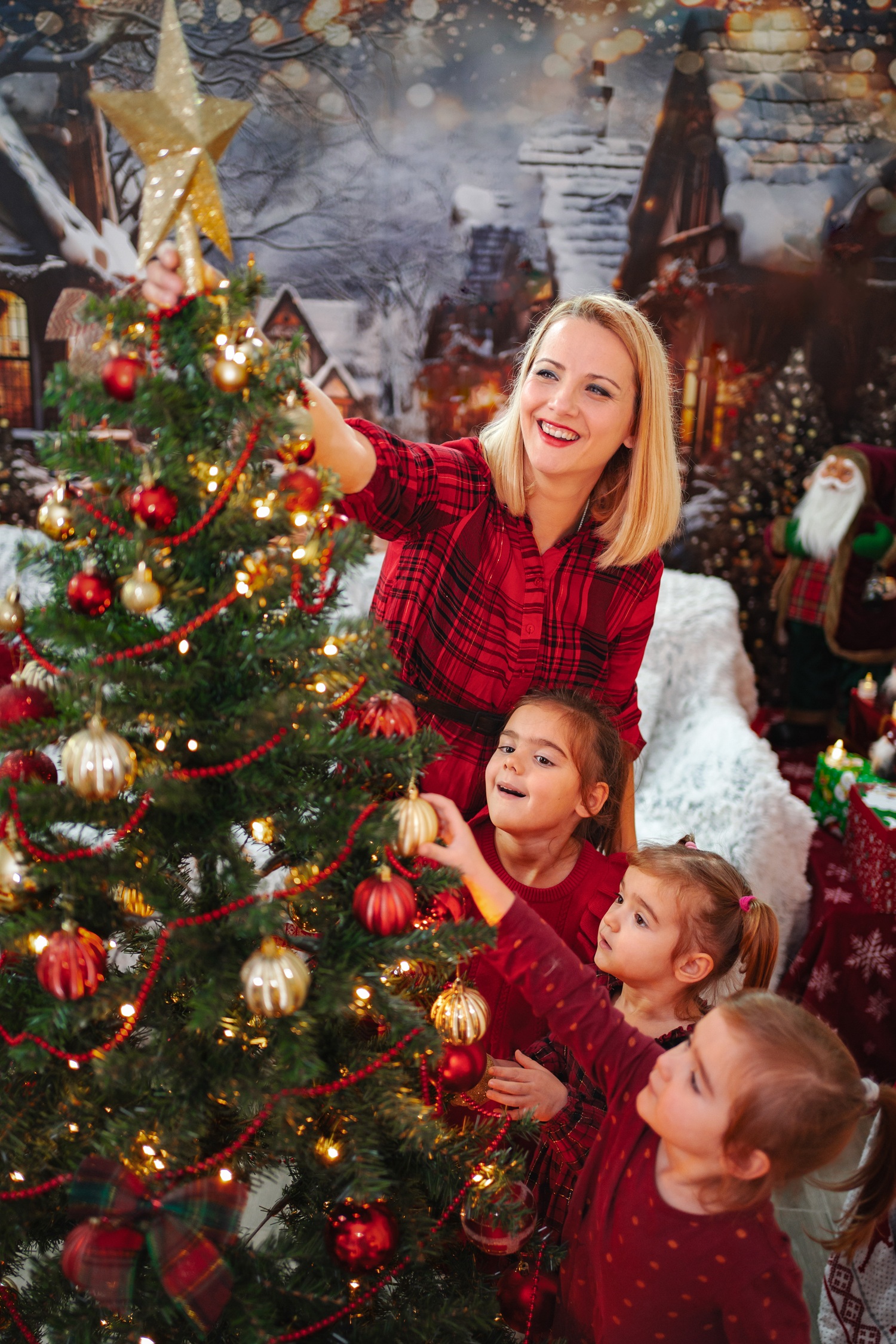 A woman and three young girls decorate a Christmas tree with ornaments and lights in a festive, holiday-themed room.