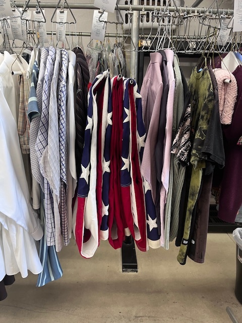 A row of hanging shirts is displayed on a rack next to several folded American flags in a dry cleaning facility.